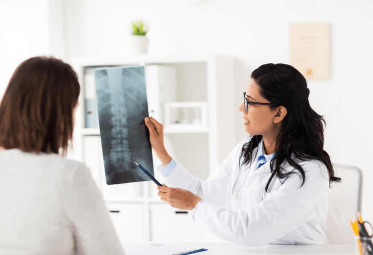 doctor in white coat and glasses pointing to an xray of a spine with a patient sitting next to her