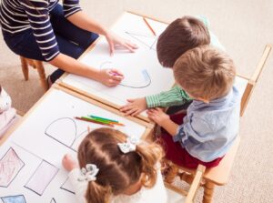 A group of young children, guided by an adult, enjoy a creative drawing session with crayons, working on large sheets of paper filled with shapes and letters.
