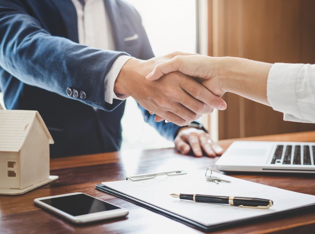 Two people shake hands over a desk with a model house, keys, and paperwork, finalizing a real estate deal in a professional setting.