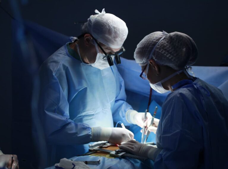 Two surgeons in an operating room, dressed in blue scrubs and surgical caps, focus intently on a procedure under bright lights.