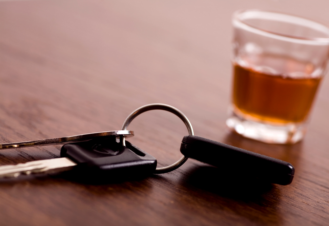 photo of car keys in the foreground and a full shot glass in the background suggesting drunk driving