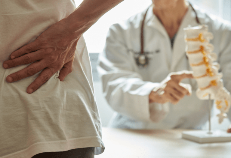 a man holds his back indicating pain while a doctor points to a spine replica