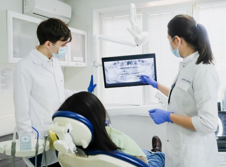 Two dental professionals in a clinic review a patient's dental X-ray on a screen while the patient sits in the examination chair.