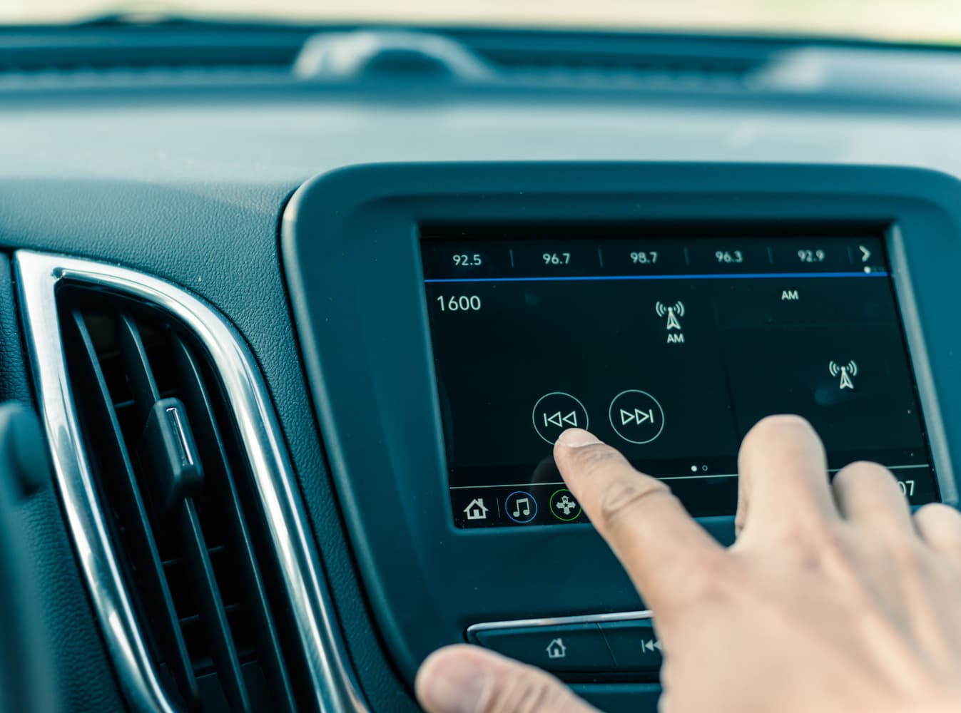 A hand adjusts the car's infotainment system, selecting the next track on the touchscreen display while tuned to AM radio at 1600 kHz.