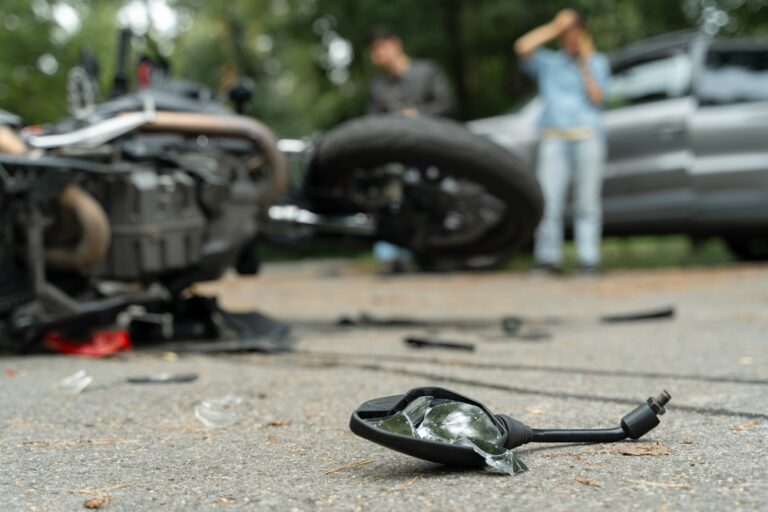 Close-up of shattered mirror with overturned motorcycle and drivers in background
