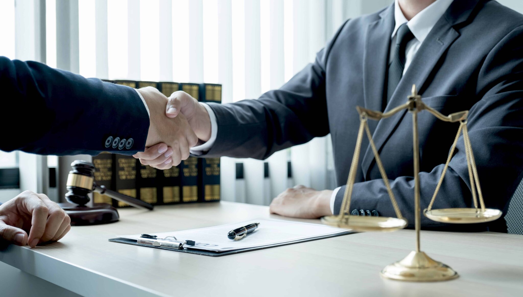 Lawyer and client shaking hands during a legal consultation with scales of justice and gavel on the desk.