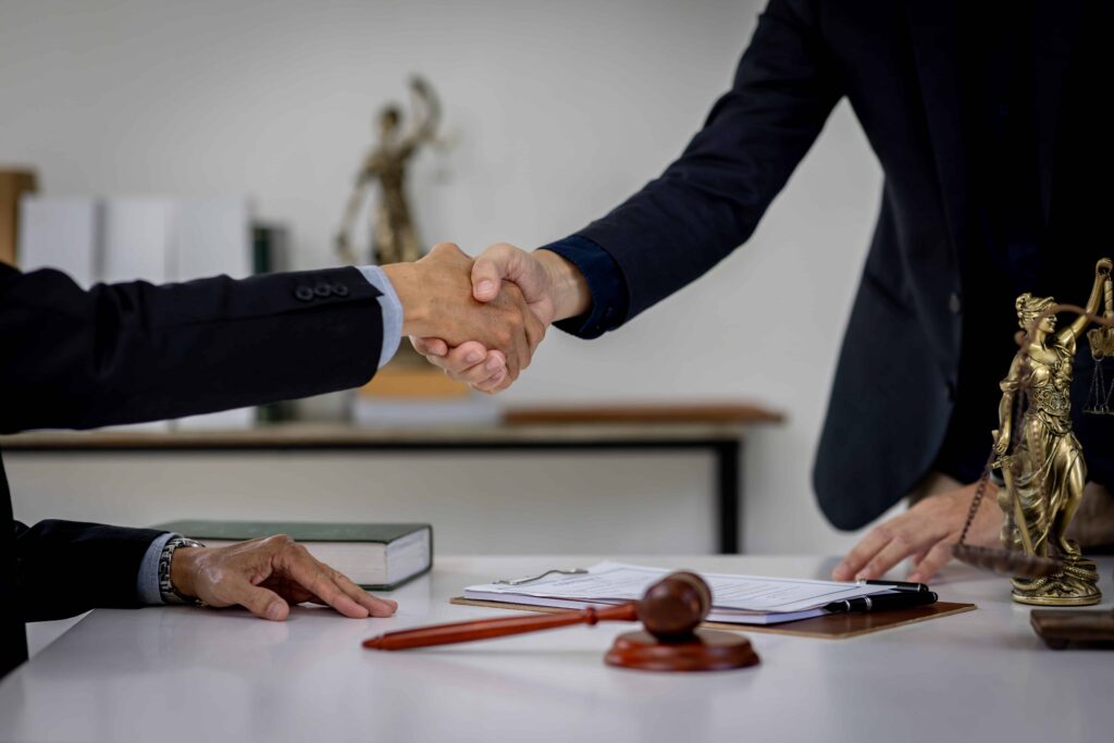 Lawyer and client shaking hands over legal documents with gavel and Lady Justice statue on desk.