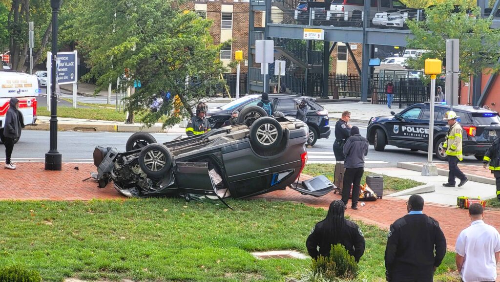 Overturned car accident scene with police, firefighters, and emergency responders assisting at an urban intersection.